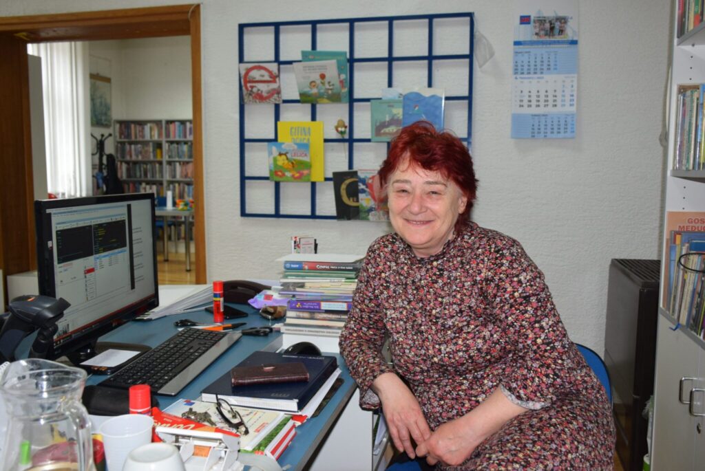 Mirjana Štelcer nasmiješena za radnim stolom u knjižnici, okružena knjigama i uredskim materijalom, u pozadini se vidi dio knjižnog fonda i dječji kutak
