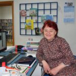 Mirjana Štelcer nasmiješena za radnim stolom u knjižnici, okružena knjigama i uredskim materijalom, u pozadini se vidi dio knjižnog fonda i dječji kutak